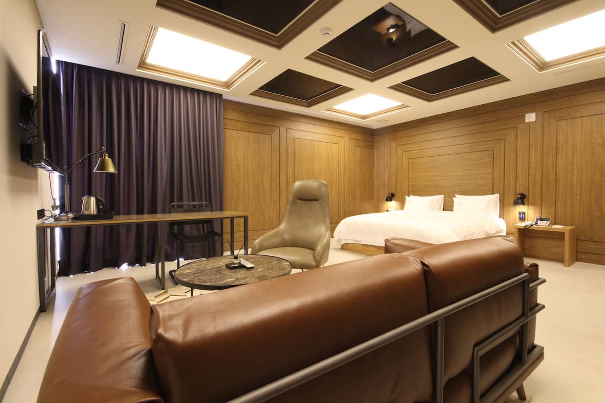 A modern hotel room with a large bed, a leather sofa, a desk with a lamp, and a TV mounted on the wall.