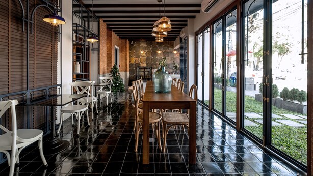 Un restaurant avec une longue table en bois et des chaises blanches, de grandes fenêtres et des lampes suspendues.