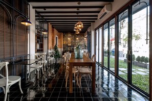Un restaurante con una larga mesa de madera y sillas blancas, ventanas amplias y lámparas colgantes.