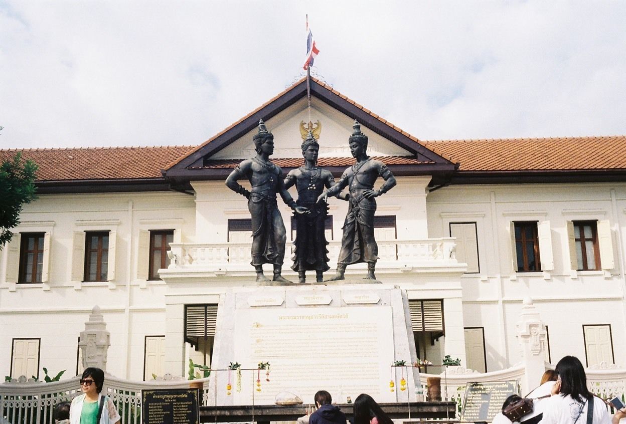 A building with a white facade, a red-tiled roof, and a statue of three figures on a pedestal.