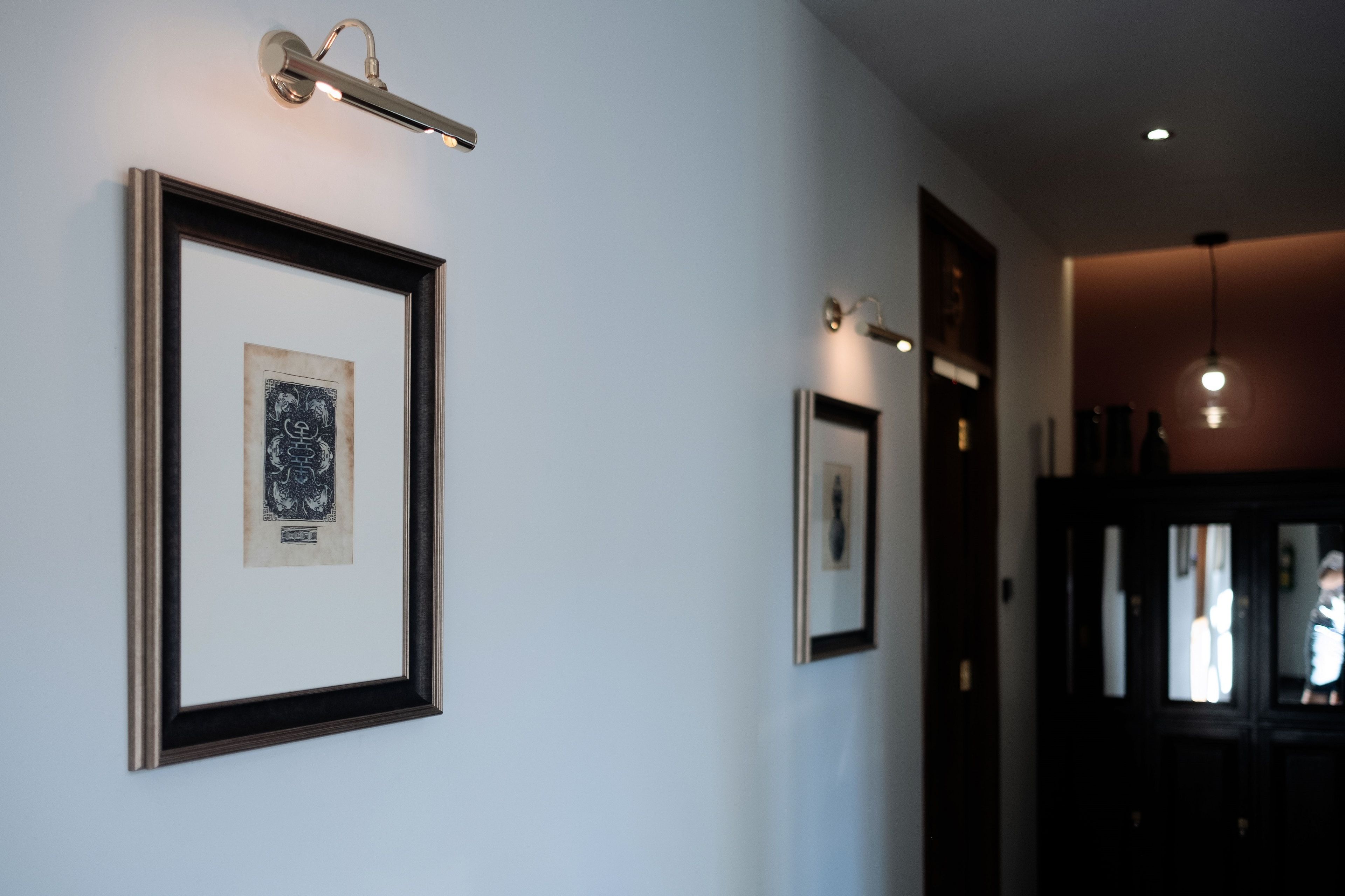 A hallway with framed artwork on the walls, a light fixture, and a door leading to another room.