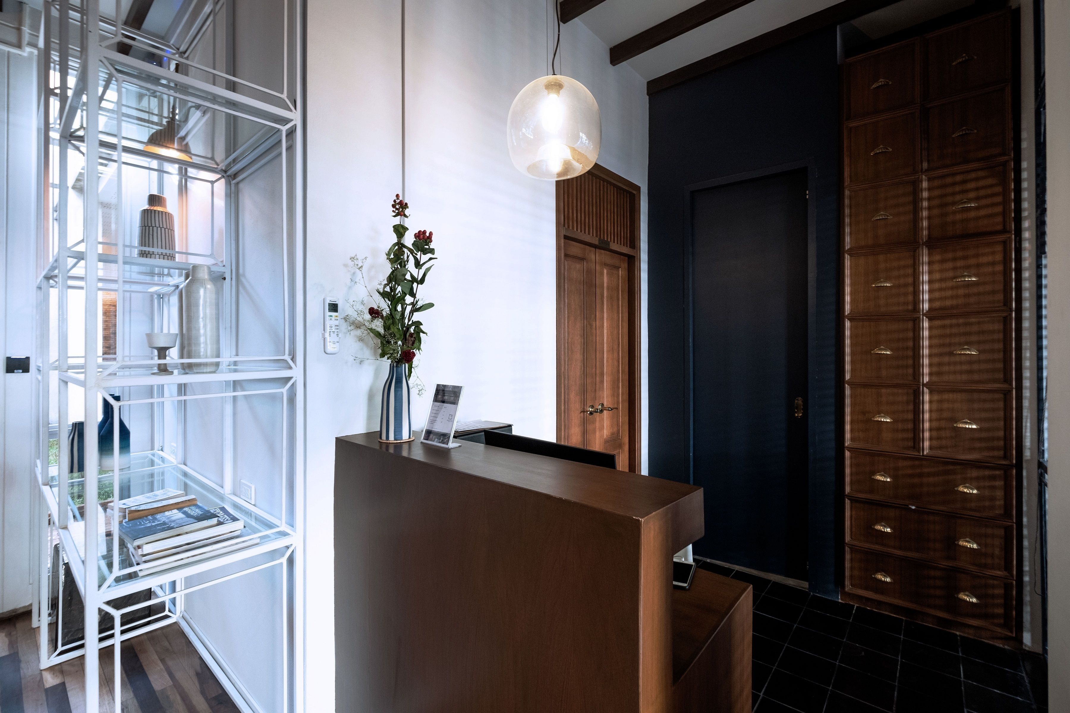 A reception area with a wooden reception desk, a glass shelving unit with books and decorative items, a hanging pendant light, and a wooden wardrobe.