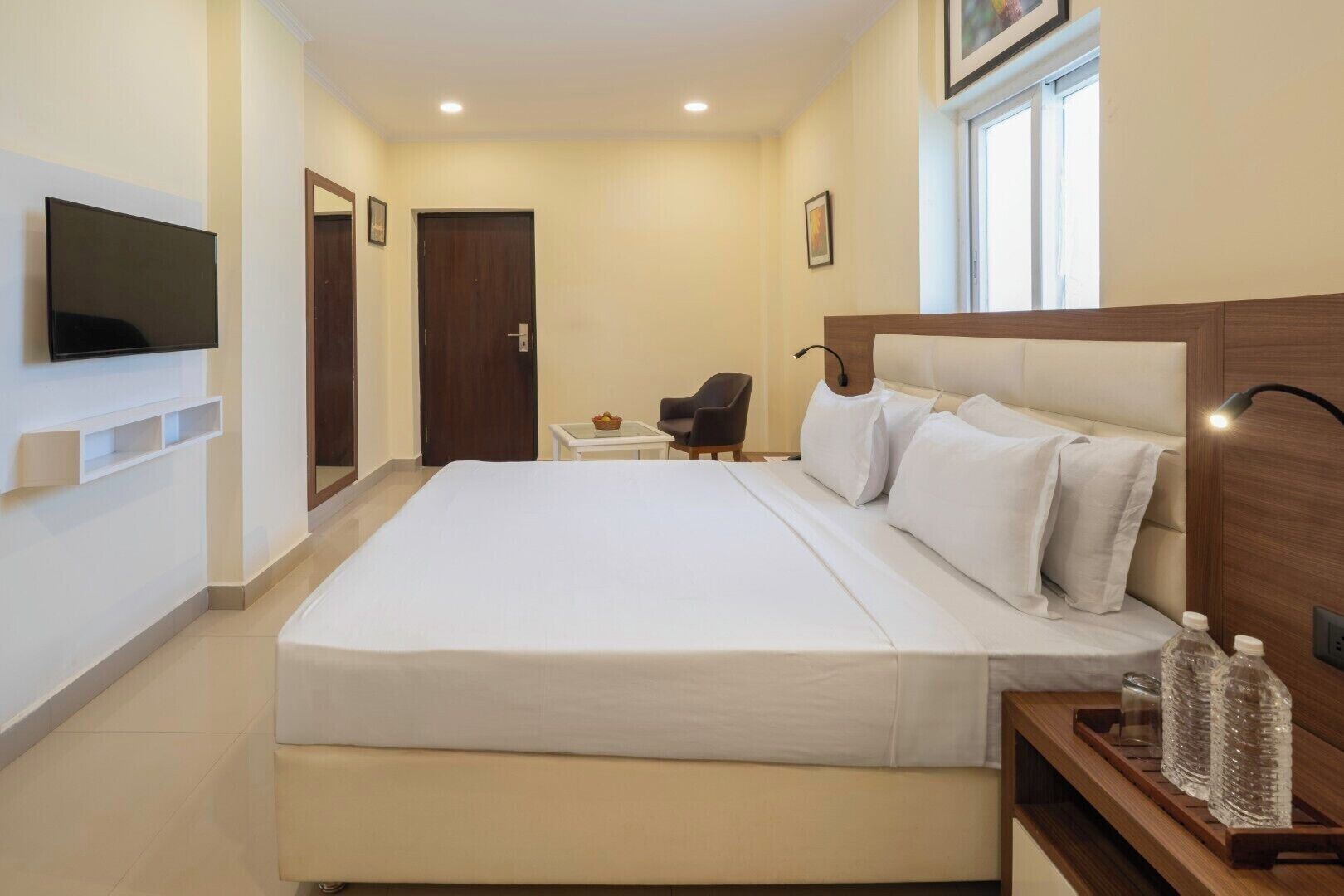 A hotel room with a large bed, a TV mounted on the wall, a small table with a chair, and a window with natural light.