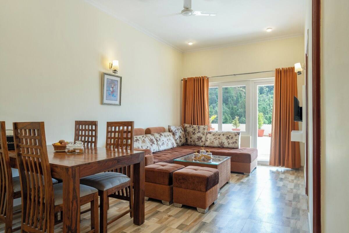 A dining area with a wooden table and chairs, a sofa with floral cushions, and a view of greenery outside.