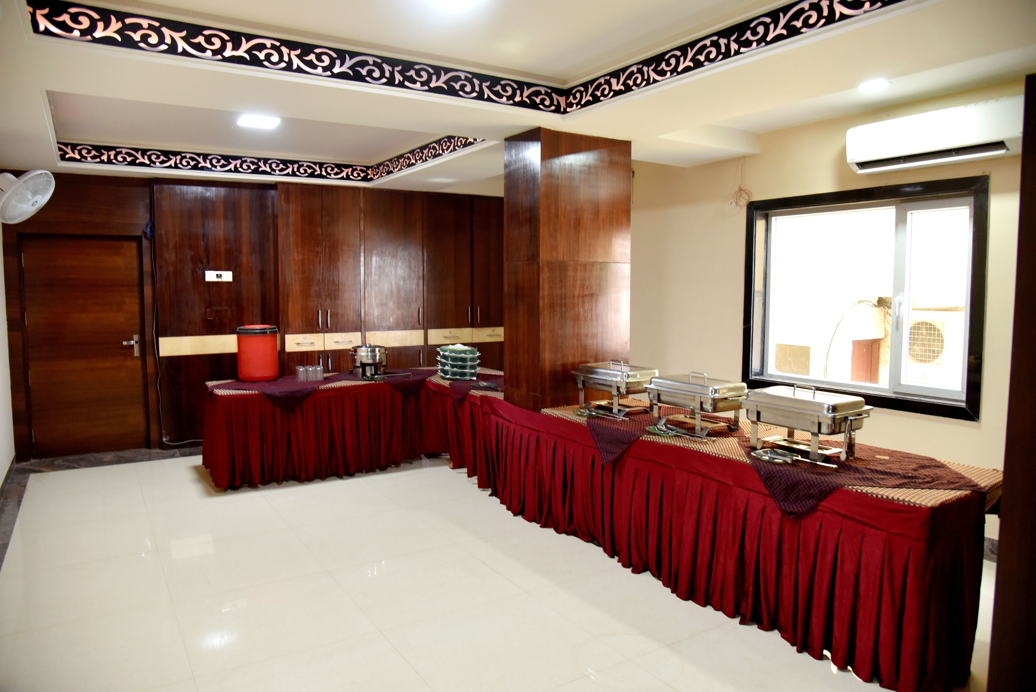 A conference room with a long table covered in a red cloth, wooden cabinets, and a window with blinds.