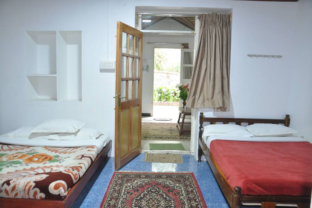 A room with two beds, a wooden door leading outside, a small table with a chair, and a red rug on the floor.