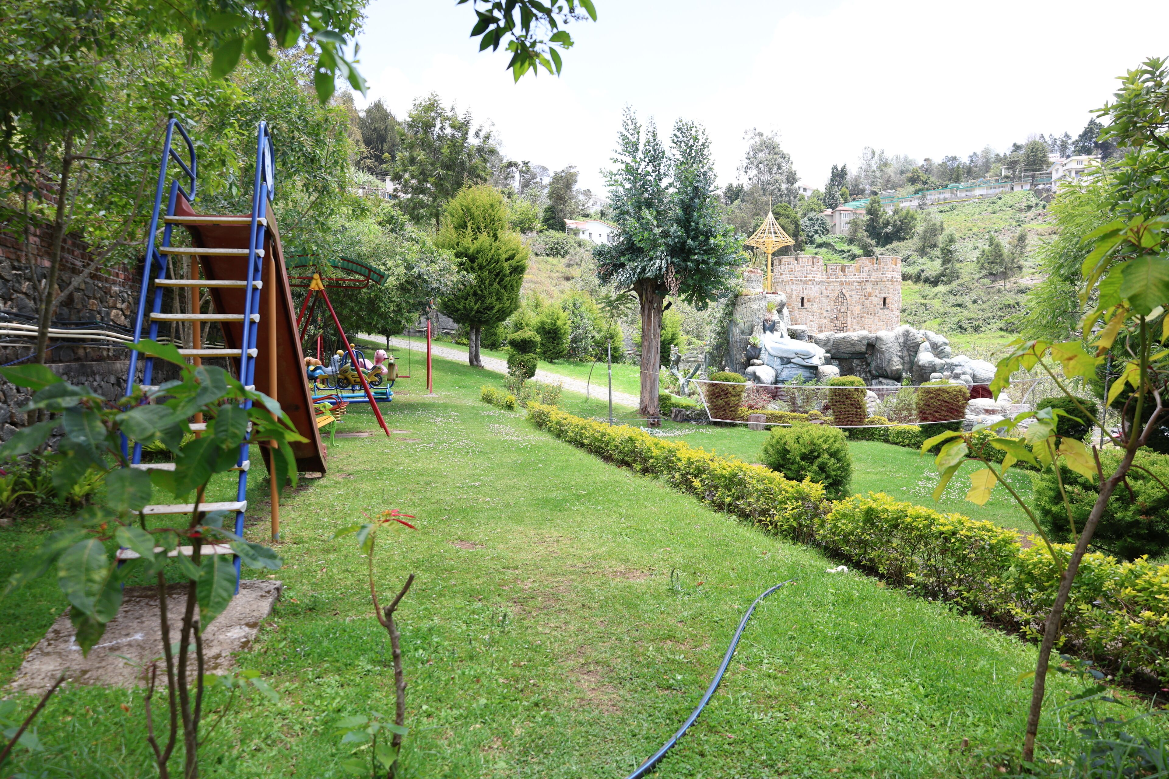 Un jardín bien cuidado con una zona de juegos infantiles, setos ordenados y una estructura de piedra que parece un castillo.