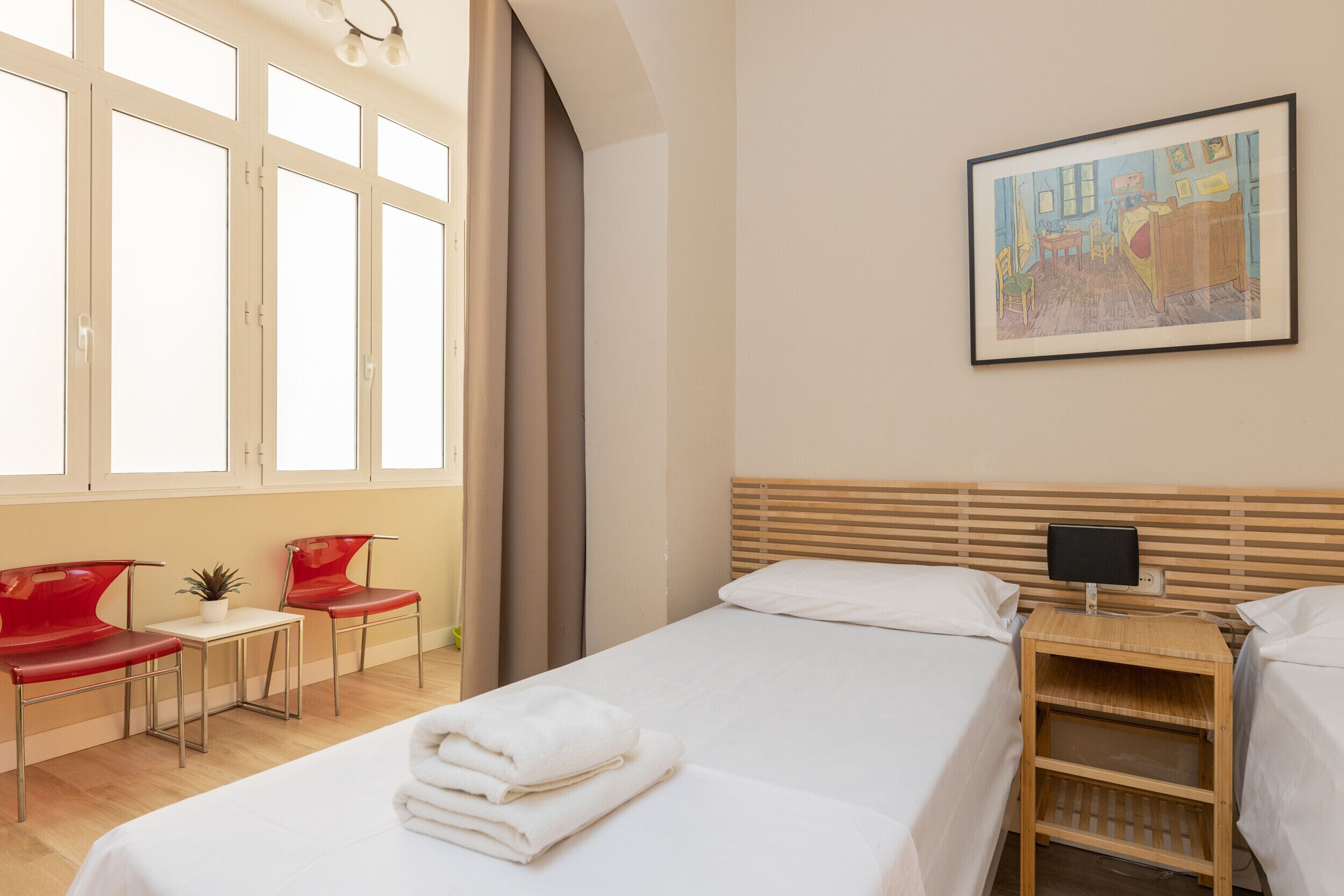 A hotel room with a bed, two red chairs, a small table with a plant, a painting on the wall, and a TV on a wooden stand.