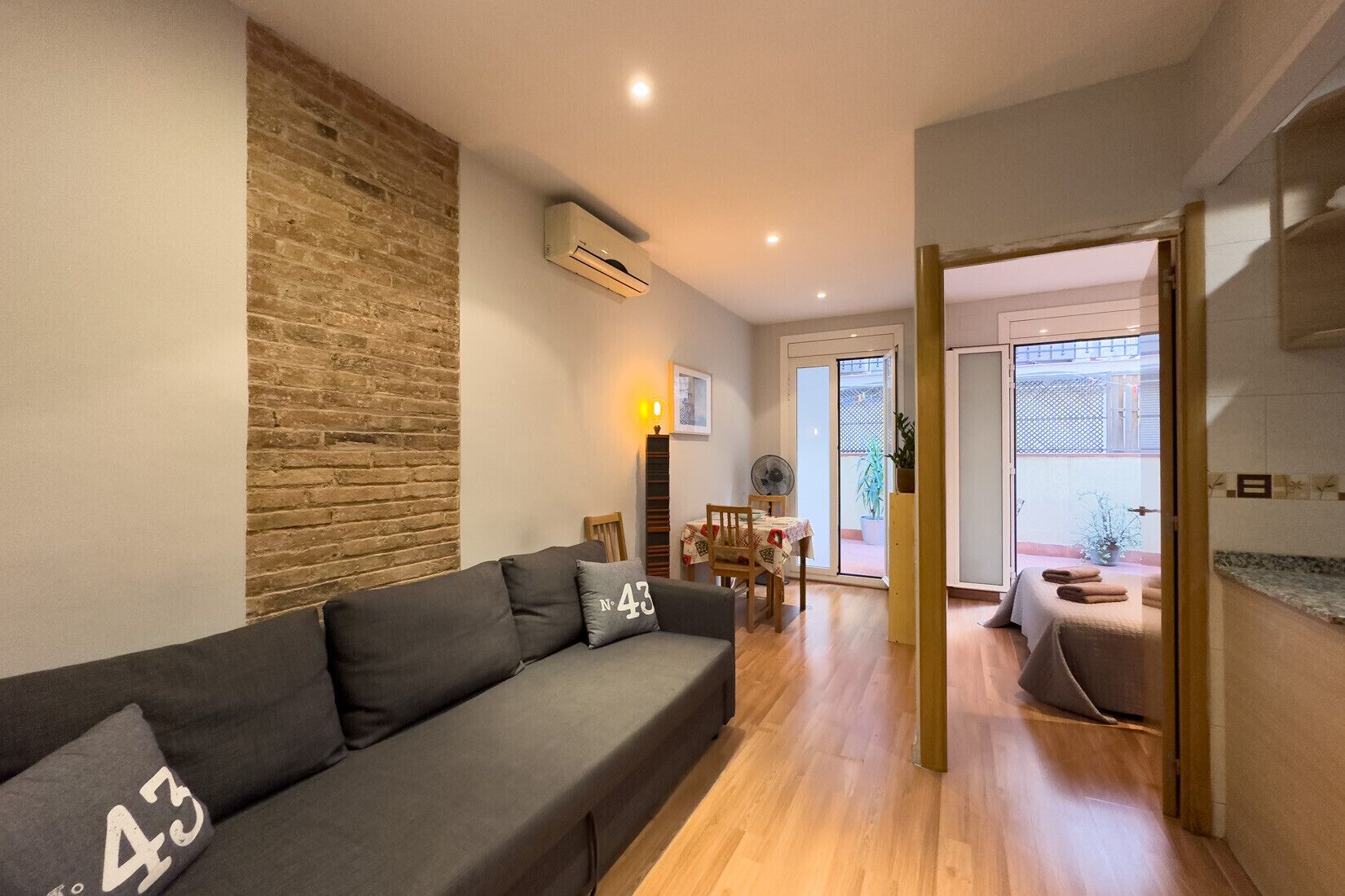 A modern living room with a grey sofa, a brick accent wall, a dining area with a table and chairs, and a sliding glass door leading to an outdoor space.