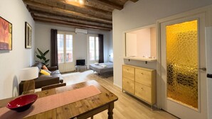 Ein gemütliches Wohnzimmer mit einem Holztisch, einer Couch und einem Fernseher. Ein Fenster mit Vorhängen, eine Pflanze und eine Kommode mit Spiegel sind ebenfalls vorhanden.