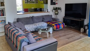 Un salon moderne avec un canapé d’angle gris, une table basse et un téléviseur. On y trouve également un coin cuisine avec des murs verts et des luminaires suspendus.