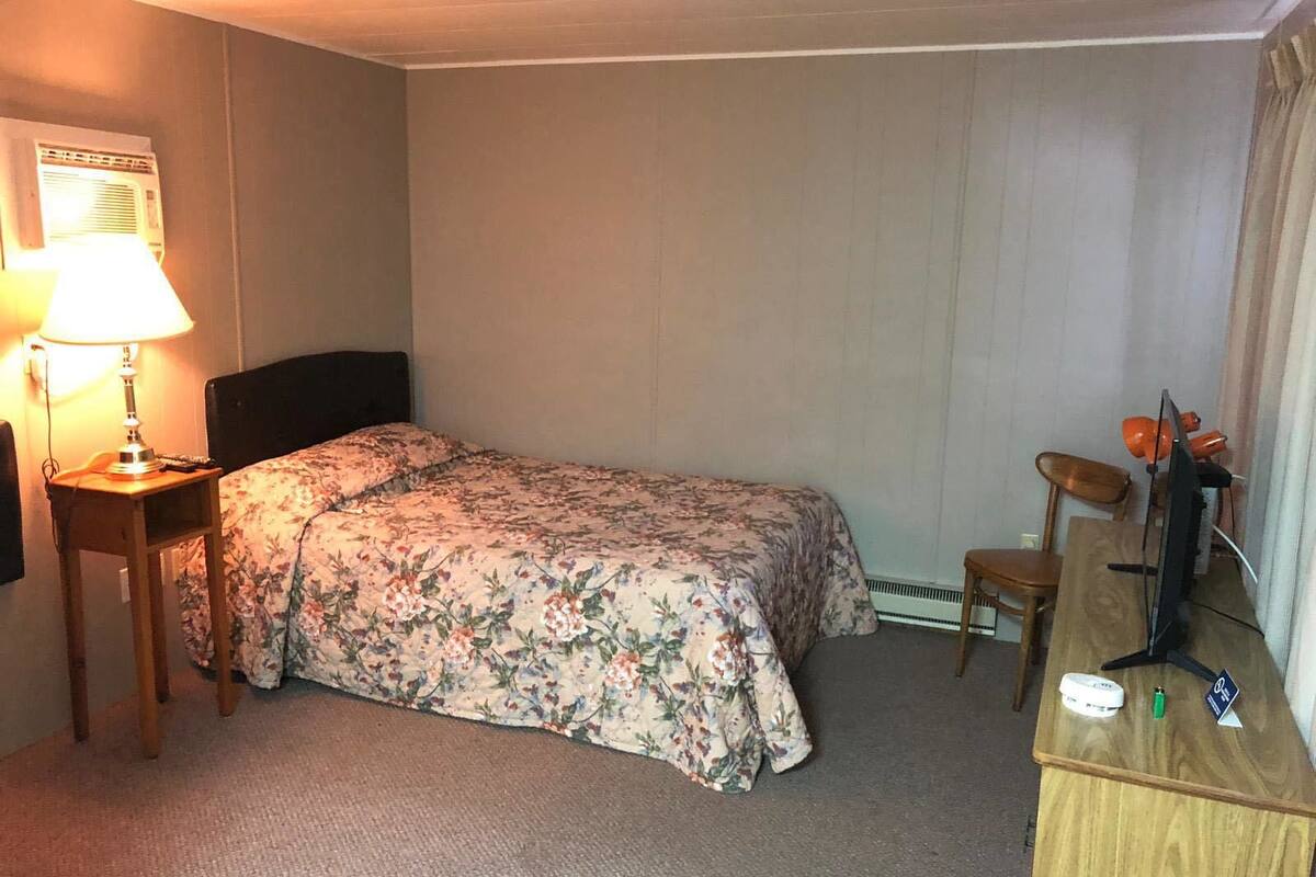 A small, neatly arranged hotel room with a single bed, a desk, a chair, and a lamp.