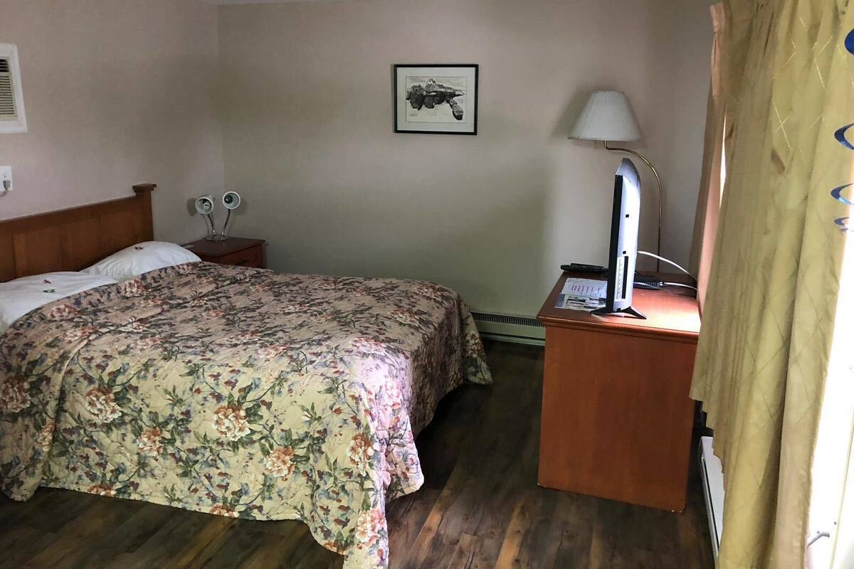 A hotel room with a bed, a desk with a computer, a lamp, a framed picture on the wall, and a window with curtains.