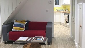 A modern living room with a grey sofa, a red throw blanket, and a brown ottoman. A kitchen is visible in the background with white cabinets and a yellow backsplash.
