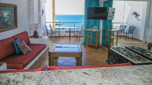 Una sala de estar con un sofá rojo, una mesa de centro de madera y un televisor sobre un mueble. Hay un balcón con vista al océano.