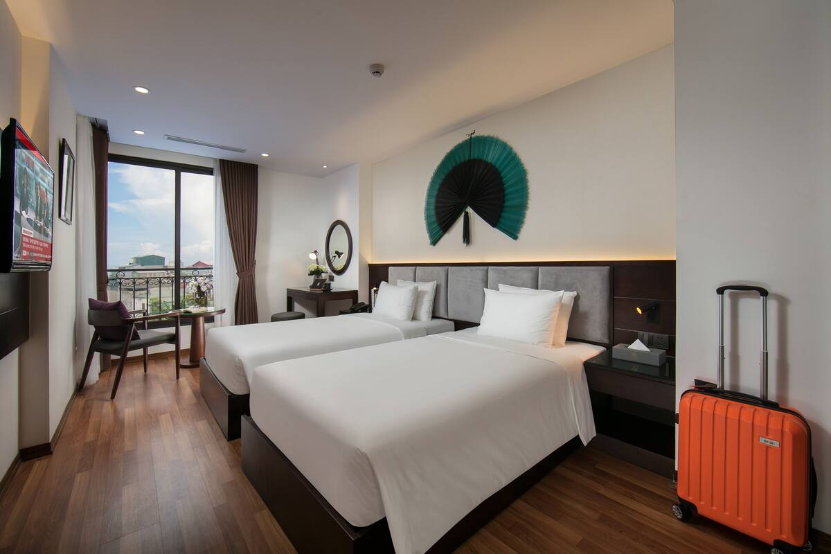 A hotel room with two beds, a desk, a chair, a large window, a wall fan, and a suitcase.