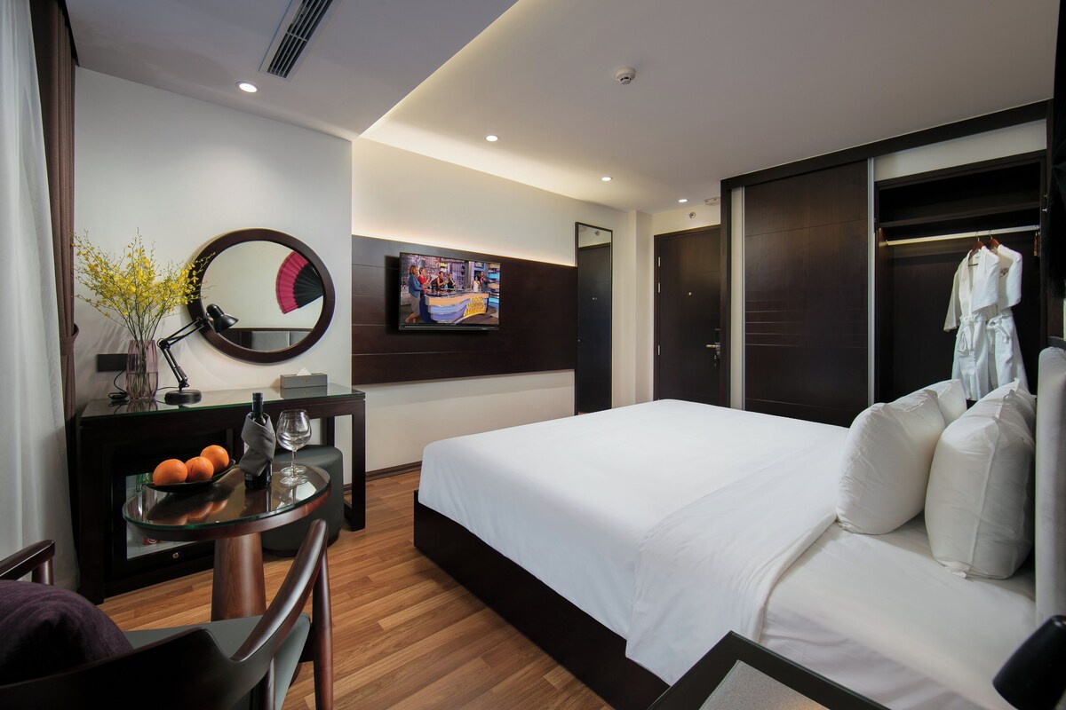 A modern hotel room with a large bed, a TV mounted on the wall, a desk with a vase of flowers, and a wardrobe.