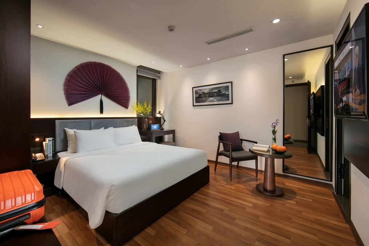 A modern hotel room with a large bed, a desk, a chair, a fan, and a mirror.