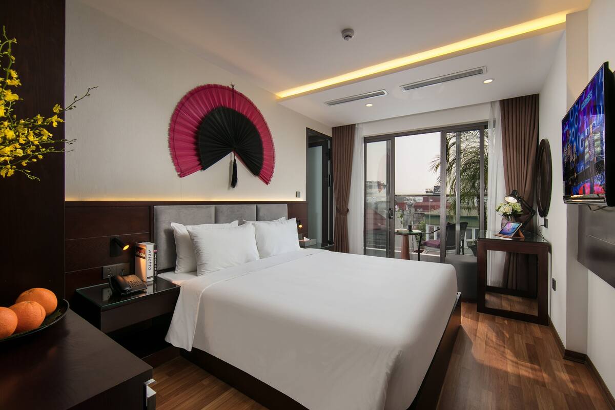 A modern hotel room with a large bed, a bedside table with a lamp, a TV, and a view of the city through large windows.