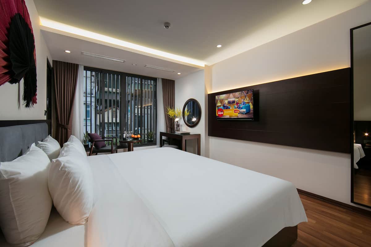 A modern hotel room with a large bed, a flat-screen TV mounted on the wall, a large window with curtains, and a view of the cityscape.