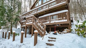 Une cabane en bois à deux étages, avec un escalier en bois menant à un balcon, entourée d’un paysage enneigé et d’arbres.