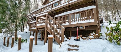 A two-story wooden cabin with a wooden staircase leading to a balcony, surrounded by snow-covered ground and trees.