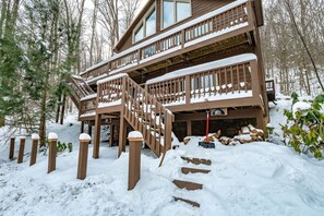 Una baita di legno a due piani con una scala in legno che conduce a un balcone, immersa in un paesaggio innevato di terra e alberi.
