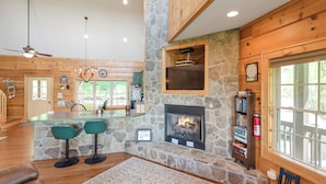 L’intérieur chaleureux d’un chalet avec une cheminée en pierre, des murs en bois, un coin repas avec des tabourets de bar et une grande fenêtre.