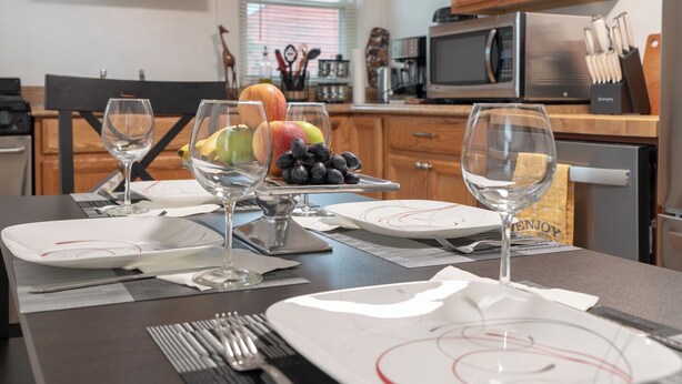 A dining table set with plates, cutlery, and glasses, with a bowl of fruit and a wine glass in the background.