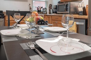 Una mesa de comedor dispuesta con platos, cubiertos y vasos, con un bol de fruta y una copa de vino al fondo.