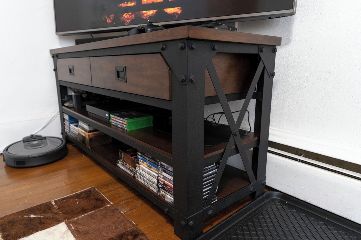 A wooden media console with shelves for books and electronic devices, a vacuum cleaner on the floor, and a television mounted above.