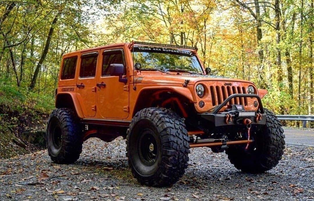 Une Jeep Wrangler, équipée de pneus tout-terrain de grande taille, est garée sur un chemin de gravier, entourée d’arbres.