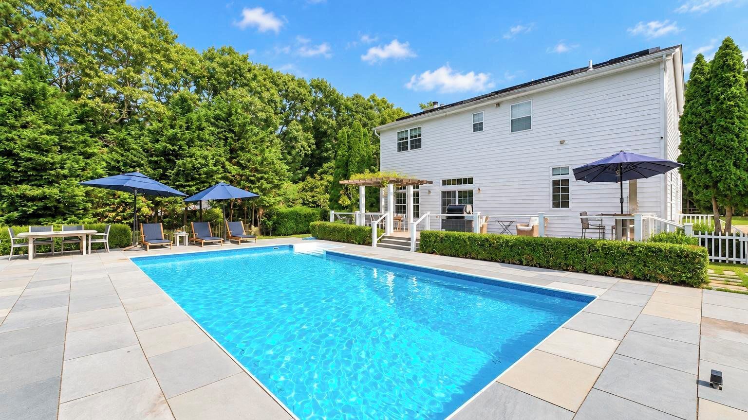 A rectangular swimming pool with a pergola and lounge chairs, surrounded by a paved patio and a white building with a covered porch.