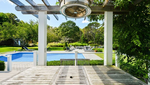Um deck de madeira com piscina, cadeiras de descanso e uma pérgula com ventilador de teto.