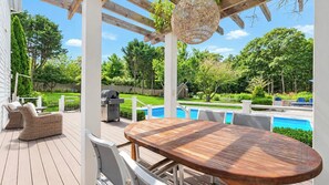 An outdoor deck with a wooden table and wicker chairs, overlooking a swimming pool and lush greenery. - Secluded Wainscott Estate with Heated Pool, Hot Tub, Expansive Yard &... (Wainscott)