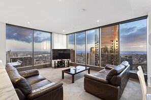 A modern living room with leather sofas, a flat-screen TV, and a coffee table. Large windows offer a city view. - Luxury Southbank Sub-Penthouse – Stunning Views, Steps to Dining & CBD (Southbank)