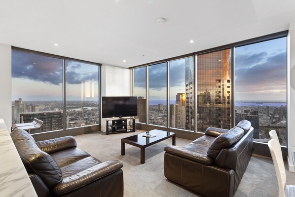 A modern living room with leather sofas, a flat-screen TV, and a coffee table. Large windows offer a city view. - Luxury Southbank Sub-Penthouse – Stunning Views, Steps to Dining & CBD (Southbank)