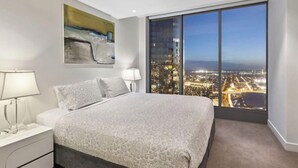 Une chambre à coucher avec un grand lit, une table de chevet, une lampe, un tableau et une vue sur la ville par une grande fenêtre.