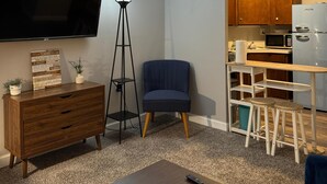 A compact living space with a flat-screen TV, a blue armchair, a wooden dresser, a floor lamp, and a kitchenette with a microwave and refrigerator. - The Westmoor (Toledo)