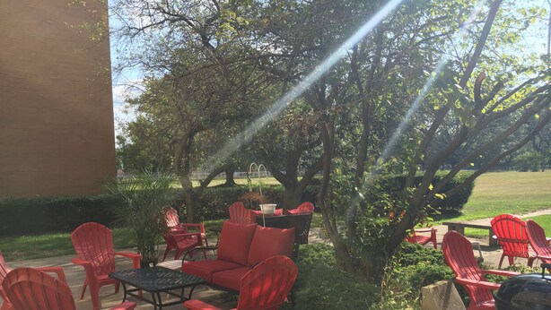 An outdoor patio with red chairs and a table, surrounded by greenery and a barbecue grill. - The Westmoor (Toledo)