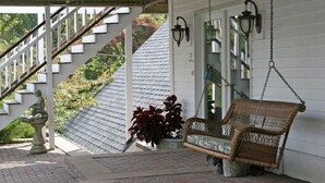 Une véranda couverte avec une balançoire en osier, une statue en pierre et un escalier menant à une maison.