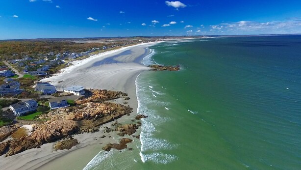 Vue aérienne d’une zone côtière avec une plage de sable, une rangée de maisons et une côte rocheuse.