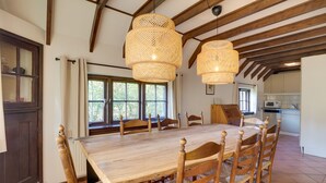 Un espace repas avec une table et des chaises en bois, de grandes fenêtres et une cuisine en arrière-plan.