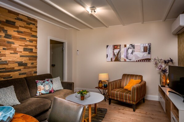 Una sala de estar moderna con una pared de acento de piedra, un sillón a rayas, un sofá, una pequeña mesa redonda con una planta y un mueble de televisión con una TV de pantalla plana.