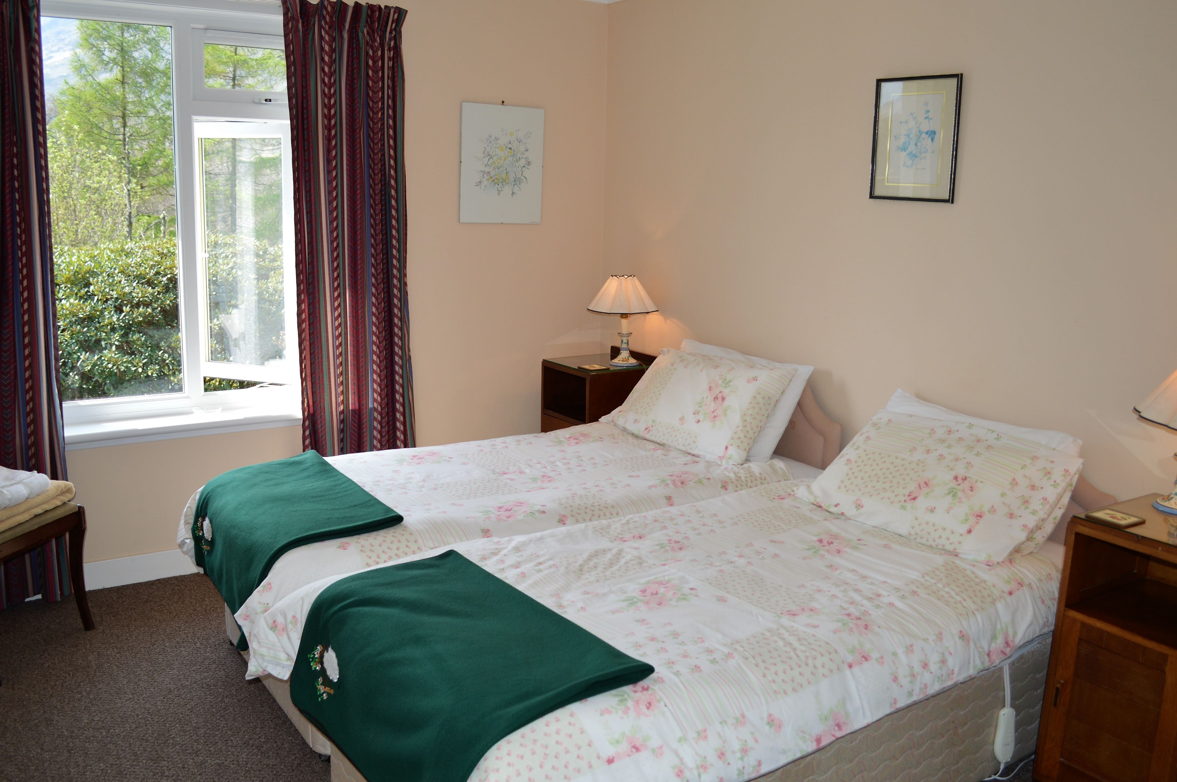 A bedroom with a bed, a nightstand, a lamp, a window with curtains, and a view of greenery outside.