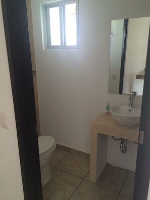 A bathroom with a toilet, a sink, a mirror, and a window. - Bungalows La Ceiba (Jiutepec)