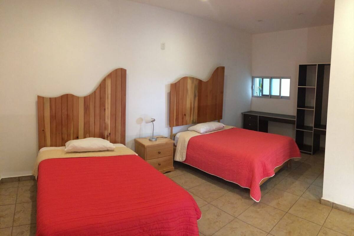 Ein Zimmer mit zwei Betten, jeweils mit einem Holz-Kopfstück, einem Nachttisch und einer Lampe. Es gibt einen Kleiderschrank und ein Fenster mit Jalousien.