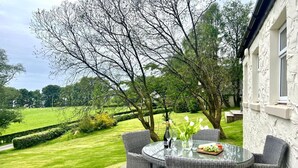 A patio with wicker furniture and a glass table, overlooking a green landscape and a white building. - Lovely cottage with loch views, near Dunoon, sleeps 6 (Blairmore)