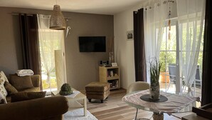 Een woonkamer met een bank, een poef en een glazen tafel met een plant. Er is een televisie aan de muur en een boekenkast.