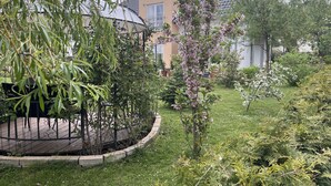Een tuin met een prieeltje, verschillende struiken en een gebouw op de achtergrond.